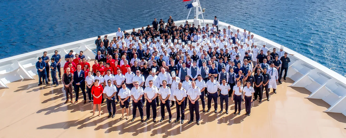 A large group of people standing on the bow of a cruise ship overlooking the water and hills in the background